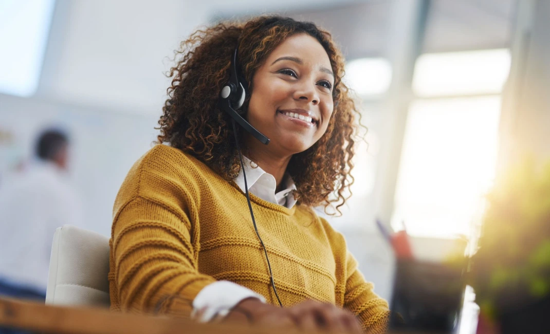 Call center agent assisting a customer on a call. Smiling woman wearing a headset while speaking with a customer in a bright call center.