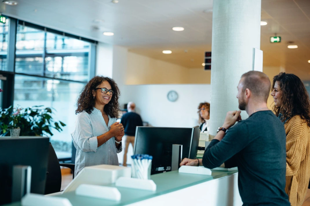 Receptionist assisting two visitors at a front desk in a modern office.